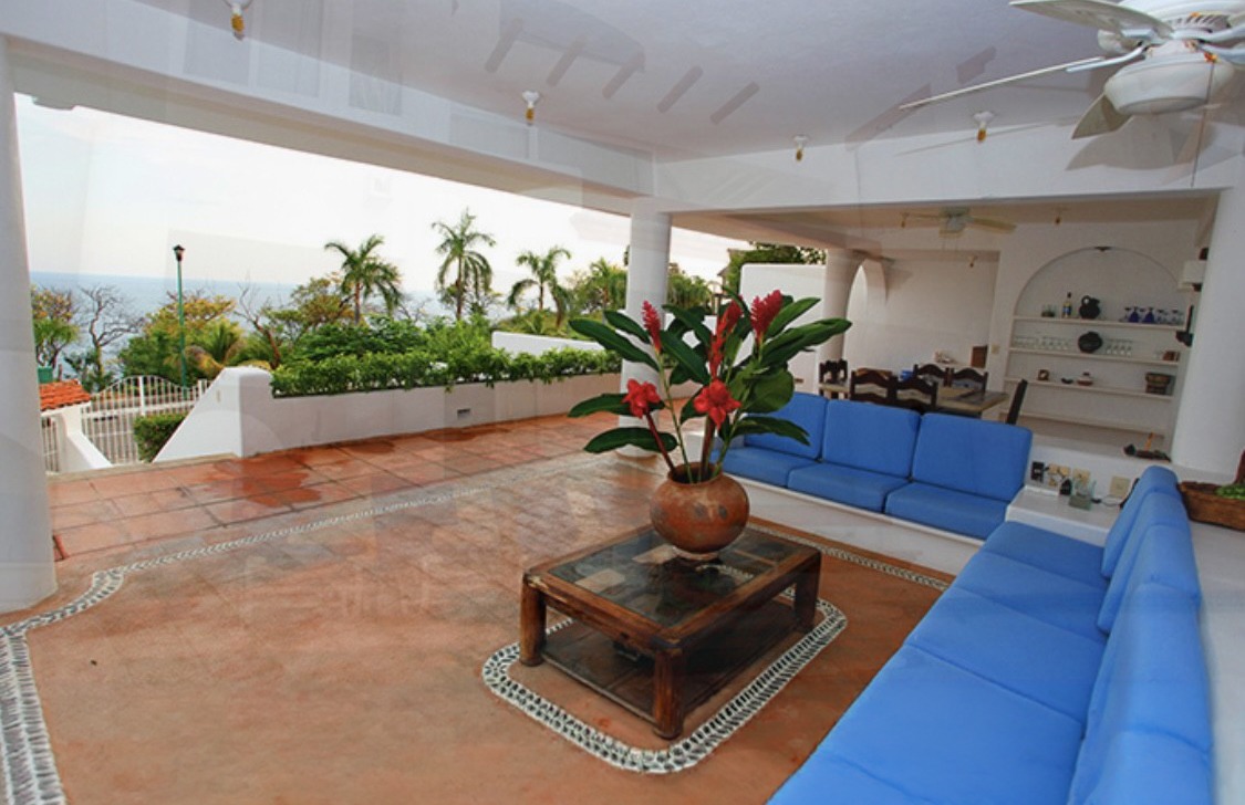 Open-air tropical patio with blue couches, tiled floors, and ocean-view greenery.