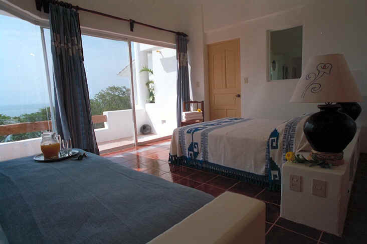 A cozy bedroom with terracotta tiles, two beds with blue and white linens, a lamp, and a tray with juice.
