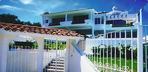 A luxurious white villa with a red-tiled roof is behind a gated entrance.