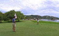 Two men on a golf course, one putting while the other holds a flag near the hole.