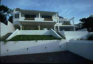Modern white two-story house with balconies, surrounded by white walls and greenery under a blue sky, conveying a sense of elegance and tranquility.