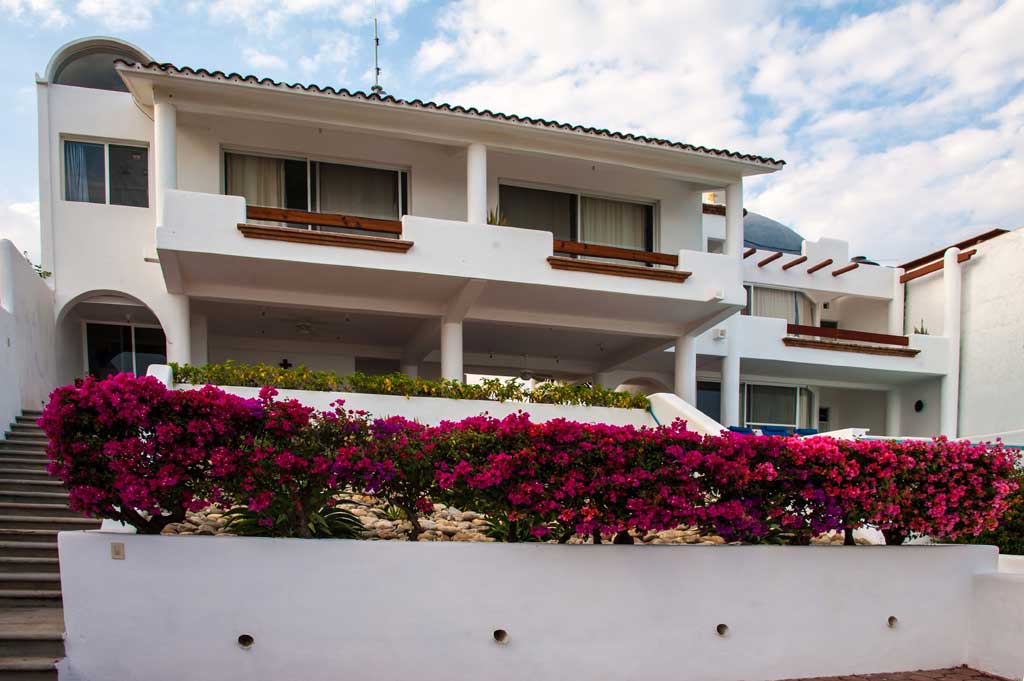 A modern, white two-story house with large windows and balconies is surrounded by vibrant pink flowers.