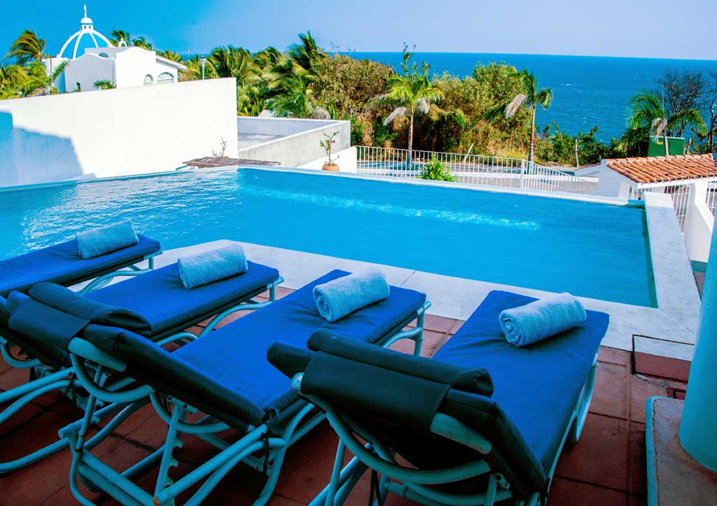 Poolside loungers with blue cushions and rolled towels face an infinity pool overlooking the ocean, surrounded by lush greenery and clear skies.