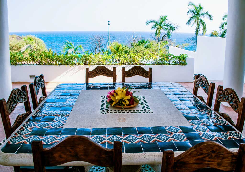 A vibrant outdoor terrace with a tiled table, surrounded by wooden chairs.